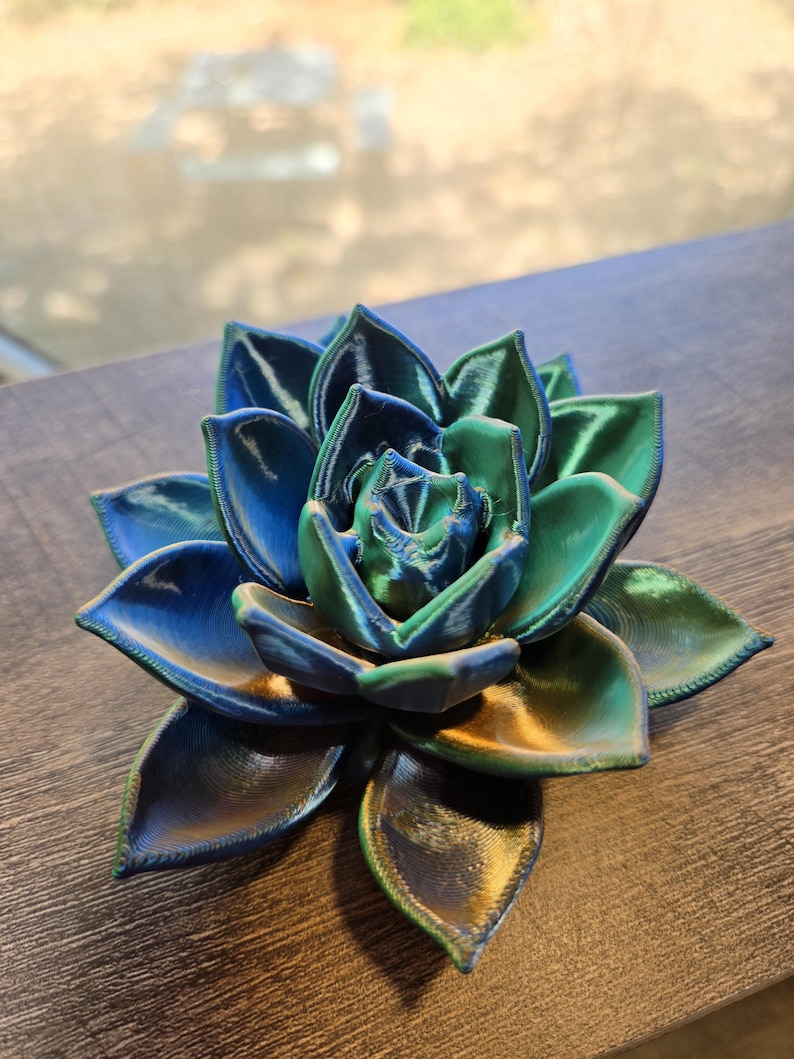 May include: A 3D printed succulent sculpture featuring layered, pointed petals in a gradient of iridescent blue, green, and gold tones. The object has a visible textured surface from the printing process, with a symmetrical, blooming rosette shape.