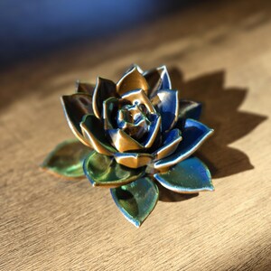 May include: A ceramic succulent sculpture featuring layered, pointed petals with a glossy finish. The color palette transitions from deep navy blue and metallic bronze at the center to vibrant emerald green and teal tones on the outer leaves.