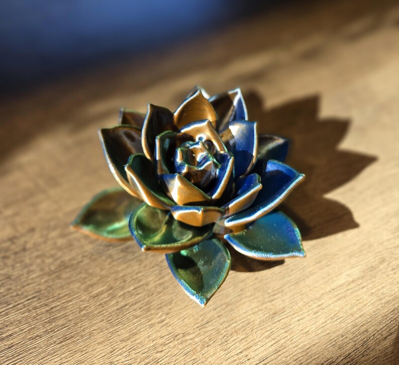 May include: A ceramic succulent sculpture featuring layered, pointed petals with a glossy finish. The color palette transitions from deep navy blue and metallic bronze at the center to vibrant emerald green and teal tones on the outer leaves.