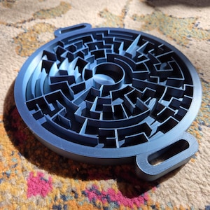 May include: A circular dark blue plastic maze puzzle featuring a complex network of raised walls and paths. The object has two small rectangular handles extending from opposite sides of the outer rim, creating a symmetrical design for the game.