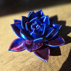 May include: A three-dimensional succulent sculpture featuring layered, pointed petals. The finish is a vibrant, glossy gradient that transitions from a deep, saturated royal blue at the center to a rich, dark purple hue at the outer petal edges.