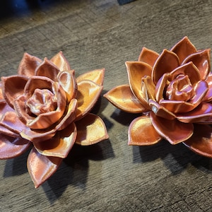 May include: Two metallic copper-colored succulent sculptures shaped like blooming flowers. Each piece features layered, pointed petals with a smooth, reflective finish that catches the light, creating a distinct sheen across the curved surfaces.