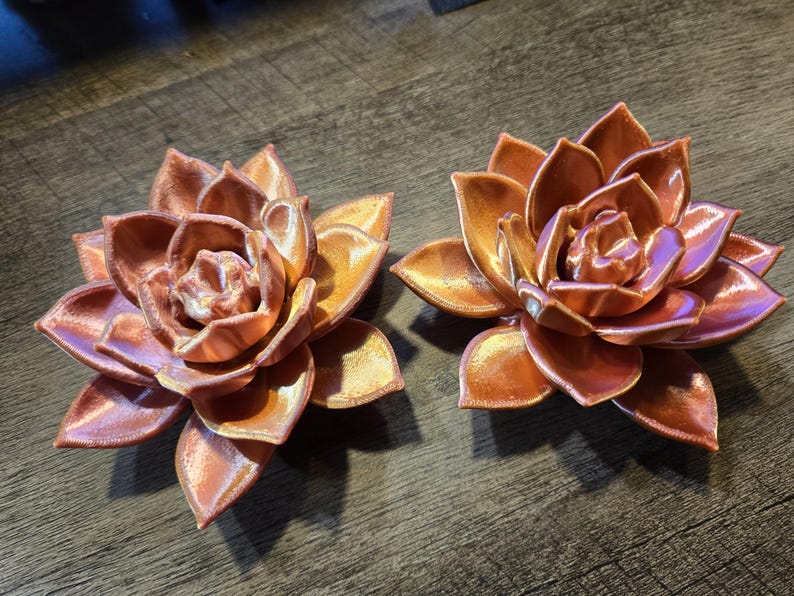 May include: Two metallic copper-colored succulent sculptures shaped like blooming flowers. Each piece features layered, pointed petals with a smooth, reflective finish that catches the light, creating a distinct sheen across the curved surfaces.
