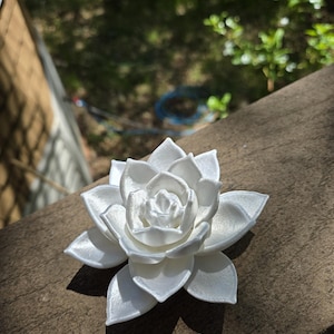 May include: A white, multi-layered succulent plant sculpture with a smooth, slightly pearlescent finish. The object features numerous overlapping, pointed petals arranged in a circular, symmetrical pattern, mimicking the appearance of a blooming flower.