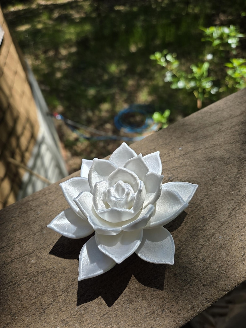 May include: A white, multi-layered succulent plant sculpture with a smooth, slightly pearlescent finish. The object features numerous overlapping, pointed petals arranged in a circular, symmetrical pattern, mimicking the appearance of a blooming flower.