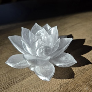 May include: A translucent white succulent figurine shaped like a blooming lotus flower. The object features layered, pointed petals with a glossy, pearlescent finish that catches the light, creating subtle highlights and shadows across its surface.