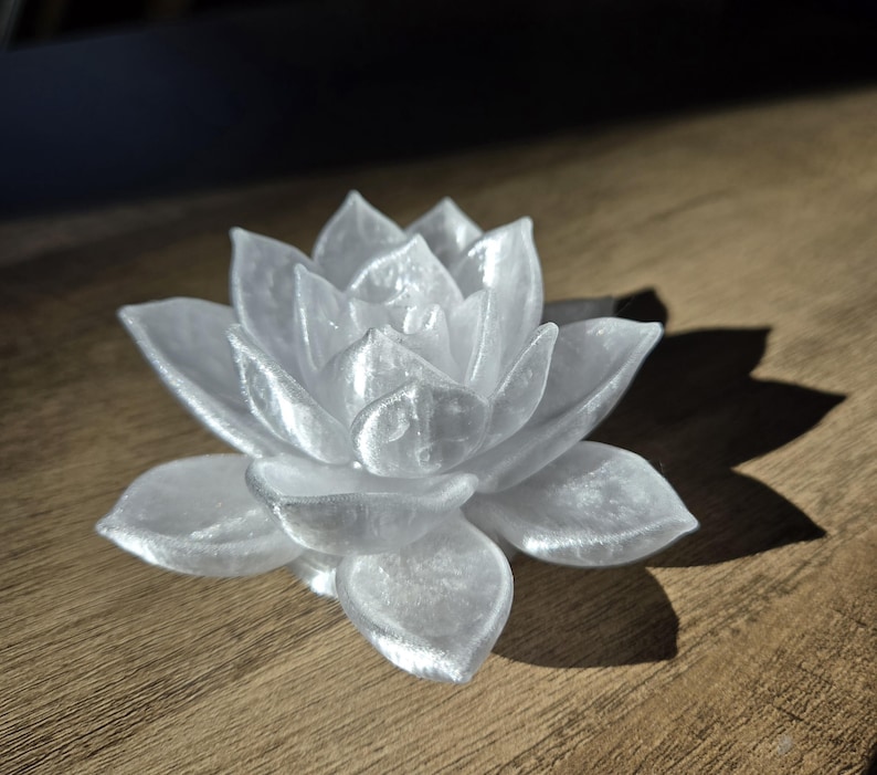 May include: A translucent white succulent figurine shaped like a blooming lotus flower. The object features layered, pointed petals with a glossy, pearlescent finish that catches the light, creating subtle highlights and shadows across its surface.