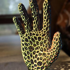 May include: A three-dimensional hand sculpture featuring an intricate, open-lattice honeycomb structure. The surface displays a gradient of metallic colors, shifting from deep green and yellow to hints of orange and purple across the mesh.