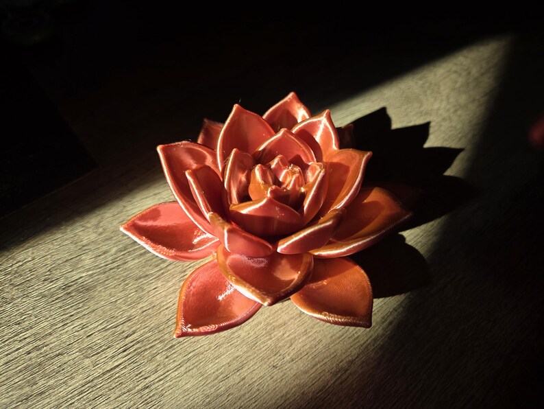 May include: A glossy, coral-colored ceramic sculpture shaped like a blooming succulent or lotus flower. The object features multiple layered, pointed petals with a smooth, reflective finish that catches the light, creating highlights along the edges.