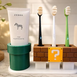 May include: A white tube of Zebra red toothpaste sits in a green cylindrical holder next to a brown brick-patterned toothbrush stand with a yellow question mark block. Three toothbrushes with black, white, and green handles are inserted.