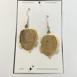 May include: A pair of dangle earrings featuring turtle designs. The earrings are made of a light brown material with etched details, and hang from silver-colored hooks. The earrings are displayed on a white card.