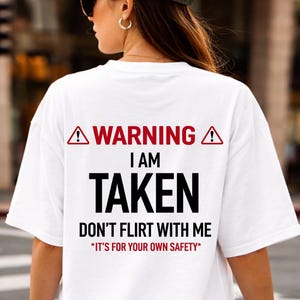 May include: White t-shirt with the text "WARNING I AM TAKEN DON'T FLIRT WITH ME *IT'S FOR YOUR OWN SAFETY" in black and red. The shirt has a relaxed fit and short sleeves. The text is centered on the back of the shirt.