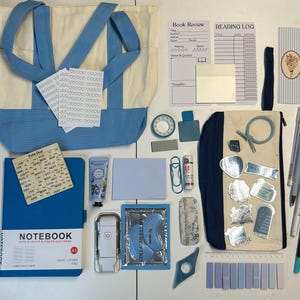 May include: A collection of blue and cream stationery items, including a tote bag, notebook, stickers, pens, and eye masks. The notebook reads "NOTEBOOK write & record & inspire your ideas". The items are arranged on a white surface.
