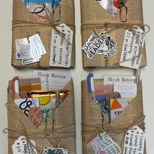 May include: Four book-themed gift packages wrapped in newspaper, tied with twine, and adorned with stickers and tags. Each package contains a pen, paper, and other small items. Tags indicate genres like suspense, romance, and fantasy.