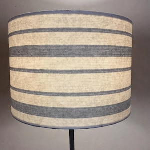 May include: A cylindrical lampshade with horizontal stripes in alternating shades of beige and blue. The lampshade has a simple, clean design, suitable for various interior styles. The base is a dark, thin metal rod.