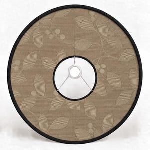 Op de afbeelding: Een ronde lampenkap met een beige stoffen buitenkant voorzien van een geweven blad- en bessenpatroon. De kap is afgewerkt met een zwarte bies langs de buiten- en binnenranden en bevat een centrale metalen spider-fitting voor montage.