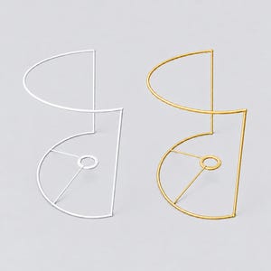 Op de afbeelding: Twee draadframes voor lampenkappen, één in een matte witte afwerking en één in een metallic gouden afwerking. Elk frame heeft een halfronde basis met een centrale ringsteun en een verticale draadstructuur die naar boven buigt in een halve cirkel.