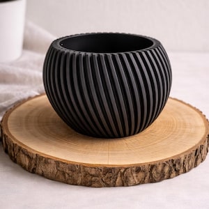 Könnte beinhalten: Ein schwarzer, kugelförmiger Pflanztopf mit einem strukturierten, geriffelten Design. Der Pflanztopf steht auf einer runden, natürlichen Holzscheibe mit einem Rindenrand. Ein weißer Topf mit einer grünen Pflanze ist im Hintergrund zu sehen.