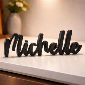 Könnte beinhalten: Ein schwarzes Holzschild mit dem Namen "Michelle" in Schreibschrift. Das Schild steht auf einer weißen Arbeitsplatte, mit einem verschwommenen Hintergrund mit einer Blumenvase.