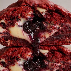 Puede incluir: Dos mitades de una galleta de terciopelo rojo apiladas. Las galletas tienen un aspecto suave y texturizado y están rellenas con un relleno cremoso blanco y una mermelada oscura similar a bayas. El relleno está goteando.