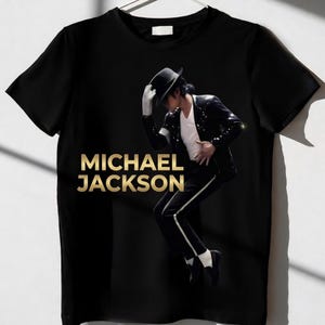Può includere: T-shirt nero con testo dorato "MICHAEL JACKSON". La maglietta mostra un'immagine di Michael Jackson in abito nero, camicia bianca, cappello nero e guanti bianchi, che esegue un passo di danza.