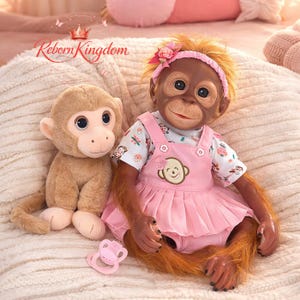 Op de afbeelding: Een levensechte babypop met bruine huid, gekleed in een roze jurk en een bloemenhoofdband, zit naast een knuffel aap. Een roze fopspeen ligt in de buurt. De pop heeft realistische kenmerken en lijkt op een baby orang-oetan.