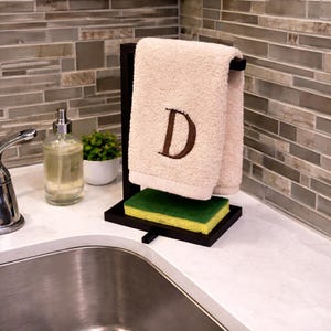 May include: A black wooden kitchen towel holder with a beige towel embroidered with the letter "D" in brown. A green and yellow sponge sits below the towel. A soap dispenser and small plant are in the background.