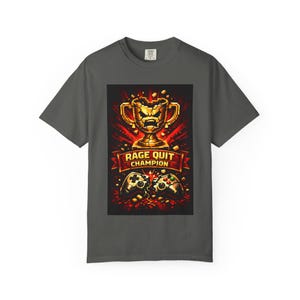 May include: Dark gray t-shirt featuring a graphic design. The design includes a pixelated gold trophy, a banner with "RAGE QUIT CHAMPION" text, and two broken game controllers. The design is in shades of red, gold, and black.