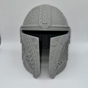 May include: A gray Mandalorian helmet with intricate tribal-style patterns. The helmet features a visor and is designed with detailed relief carvings across the surface. The overall design is symmetrical and appears to be 3D-printed.