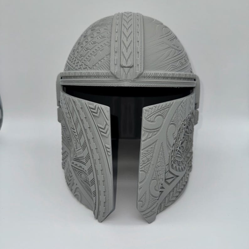 May include: A gray Mandalorian helmet with intricate tribal-style patterns. The helmet features a visor and is designed with detailed relief carvings across the surface. The overall design is symmetrical and appears to be 3D-printed.