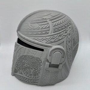 May include: A gray Mandalorian helmet with intricate tribal patterns. The helmet features a visor and detailed relief carvings across the surface, showcasing a blend of geometric and organic designs. The helmet is a solid, three-dimensional object.