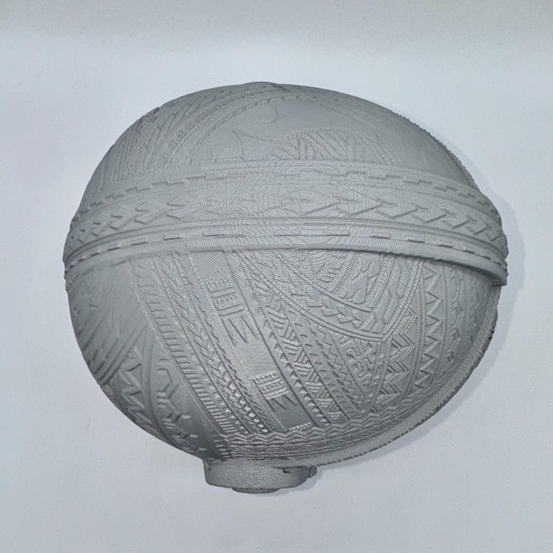 May include: A gray, spherical object with intricate, geometric patterns carved across its surface. The design includes bands of detailed textures and a small, rounded base. The overall aesthetic suggests a handcrafted or ornamental piece.