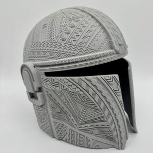May include: A gray helmet with intricate, raised tribal patterns covering the entire surface. The helmet has a visor and ear pieces, and the design is detailed with geometric shapes and lines. The helmet is likely a costume or collectible item.