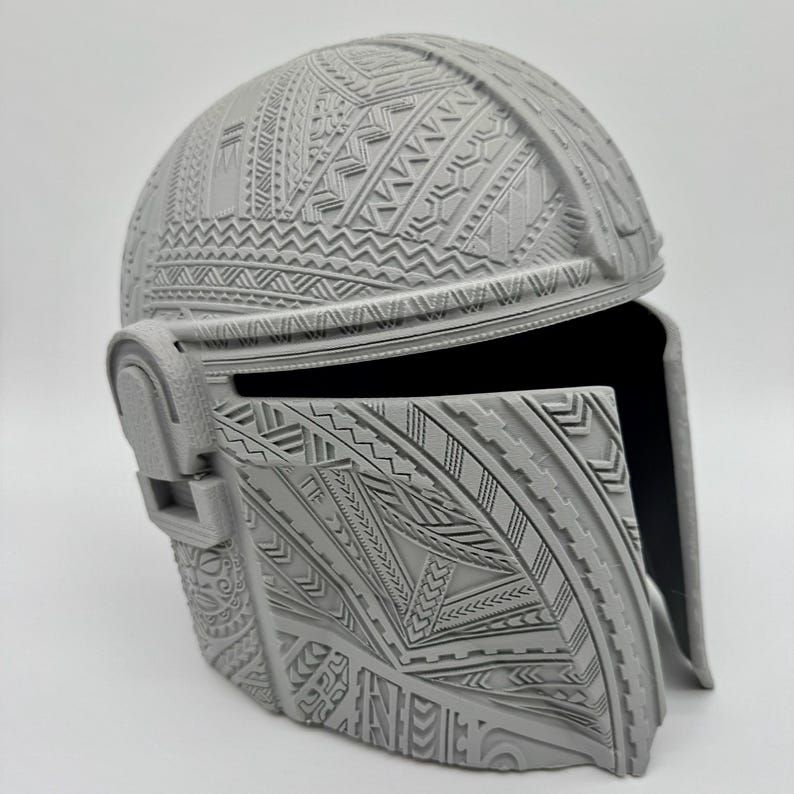 May include: A gray helmet with intricate, raised tribal patterns covering the entire surface. The helmet has a visor and ear pieces, and the design is detailed with geometric shapes and lines. The helmet is likely a costume or collectible item.