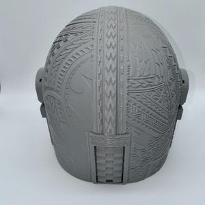 May include: A gray, intricately patterned helmet with detailed tribal designs. The helmet features a rounded shape with side attachments and a rectangular opening at the base. The surface is covered in carved geometric and curvilinear patterns.