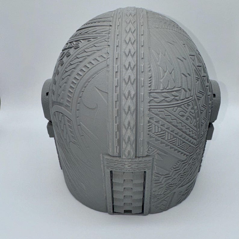 May include: A gray, intricately patterned helmet with detailed tribal designs. The helmet features a rounded shape with side attachments and a rectangular opening at the base. The surface is covered in carved geometric and curvilinear patterns.