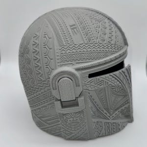 May include: A gray Mandalorian helmet with intricate tribal patterns covering the entire surface. The helmet features a visor and ear pieces, showcasing detailed relief carvings. The design is a blend of futuristic and traditional aesthetics.