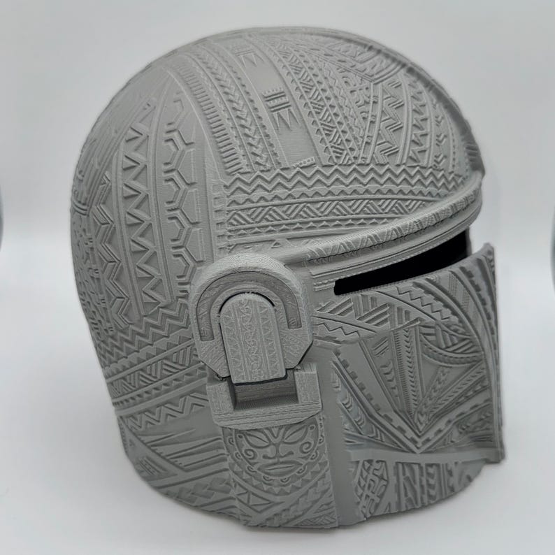 May include: A gray Mandalorian helmet with intricate tribal patterns covering the entire surface. The helmet features a visor and ear pieces, showcasing detailed relief carvings. The design is a blend of futuristic and traditional aesthetics.