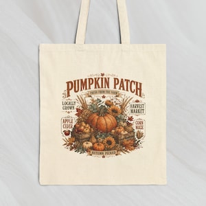 May include: A beige canvas tote bag featuring a central graphic with the text PUMPKIN PATCH, FRESH FROM THE FARM, LOCALLY GROWN, HARVEST MARKET, APPLE CIDER, CORN MAZE, and AUTUMN PICKED, surrounded by illustrations of pumpkins, sunflowers, and wheat.