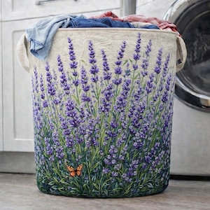 May include: A cylindrical laundry hamper with a lavender field design. The hamper features a cream-colored background with a vibrant illustration of purple lavender flowers and green stems. An orange butterfly is near the bottom. The hamper has two handles and is filled with clothes.