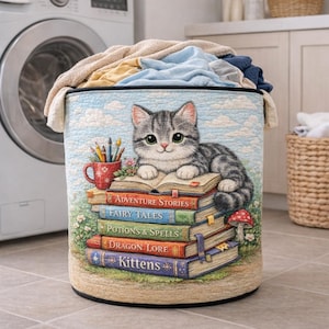 May include: A cylindrical laundry hamper with a whimsical design. The hamper features a gray tabby kitten resting on a stack of books with titles like "Adventure Stories" and "Fairy Tales." A red mug with pencils sits nearby. The hamper is filled with folded laundry.