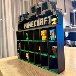 Könnte beinhalten: Ein schwarzes, 3D-gedrucktes Regal mit dem Minecraft-Logo an der Oberseite. Das Regal enthält mehrere quadratische Fächer, die jeweils mit grünem Text beschriftet sind, der verschiedene Charaktere und Gegenstände wie Alex, Steve, Creeper und andere benennt.