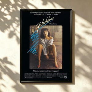 May include: Framed movie poster for the film "Flashdance." The poster features a woman in a gray top and red heels. The title "Flashdance" is in a stylized font with blue accents. The poster includes the text "Take your passion and make it happen!"