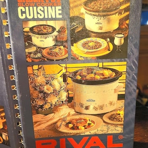 May include: A cookbook titled "Crock-Pot Slow Cooker Cuisine" with images of slow cookers and prepared dishes. The cover is blue with the brand name "RIVAL" in red letters. The cookbook is spiral-bound.