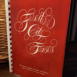 May include: A red cookbook titled "Furniture City Feasts" with white cursive lettering. The cover also includes the text "The Junior League of High Point, Inc. High Point, North Carolina 27262". The cookbook is spiral-bound.
