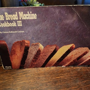 May include: A vintage cookbook titled "The Bread Machine Cookbook III" with a purple cover and images of sliced bread. The book's title is in white, with the author's name below. The cover is slightly worn, suggesting it's a well-used kitchen resource.