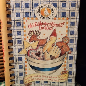 May include: A spiral-bound cookbook titled Old-Fashioned Country Cookies by Gooseberry Patch Co. The cover features a blue and white plaid pattern, a bowl filled with various shaped cookies, and the text Yummy recipes, tips, traditions, how-tos, and sweet memories... everything Cookies!