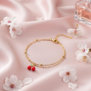 Peut inclure: Un bracelet doré à double chaîne avec un charm en forme de cerise. Le charm représente deux cerises rouges avec des feuilles vertes. Le bracelet est présenté sur une surface en satin rose, entouré de fleurs blanches.