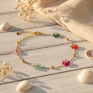 Peut inclure: Un bracelet chaîne doré orné de perles en forme de fleurs colorées. Le bracelet présente des accents de fleurs roses, vertes et rouges, avec de petites perles dorées le long de la chaîne. Le bracelet est présenté sur une surface en bois blanc.