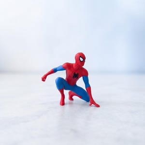 Pode incluir: Uma pequena figura do Homem-Aranha em posição agachada, com um traje vermelho e azul, um emblema de aranha preto no peito e lentes oculares brancas. O personagem está posicionado com uma mão tocando o chão e um joelho dobrado.